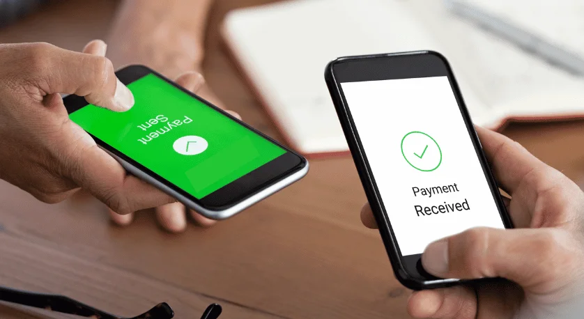 Two hands holding smartphones displaying a successful Bitcoin payment using the Lightning Network. One screen shows 'Payment Sent' with a progress indicator, and the other shows 'Payment Received' with a green checkmark. The background includes a laptop and a wooden desk.