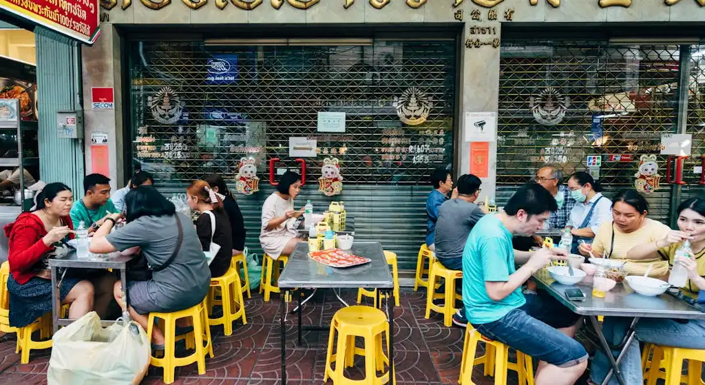 A lively street-side restaurant in Thailand, with patrons seated on yellow stools at metal tables, enjoying meals and conversations. The restaurant has a closed metal shutter adorned with decorations and signs, and the atmosphere is casual and communal.