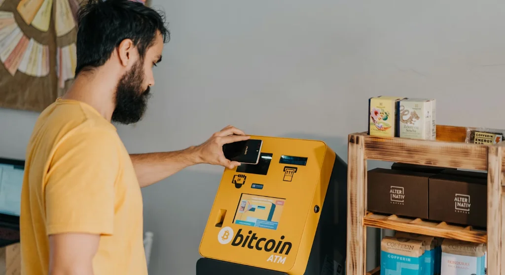 A man using a Bitcoin ATM to make a transaction, holding a smartphone in his right hand and interacting with the machine's screen. The Bitcoin ATM is yellow with the Bitcoin logo and text 'bitcoin ATM.' The setting appears to be a small shop or café with shelves of products in the background.